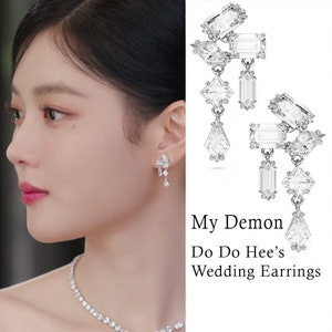 Op de afbeelding: Een close-up van een paar zilveren oorhangers met meerdere heldere kristallen. De oorhangers worden getoond aan een vrouwen oor. De tekst "My Demon Do Do Hee's Wedding Earrings" is zichtbaar onder de oorhangers.