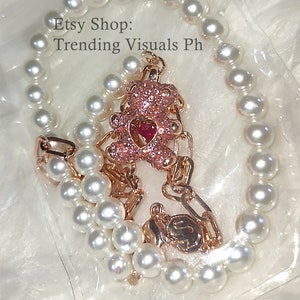May include: A gold chain necklace with a pink sparkly teddy bear charm and a white pearl bracelet. The necklace and bracelet are on a white background. Etsy Shop: Trending Visuals Ph
