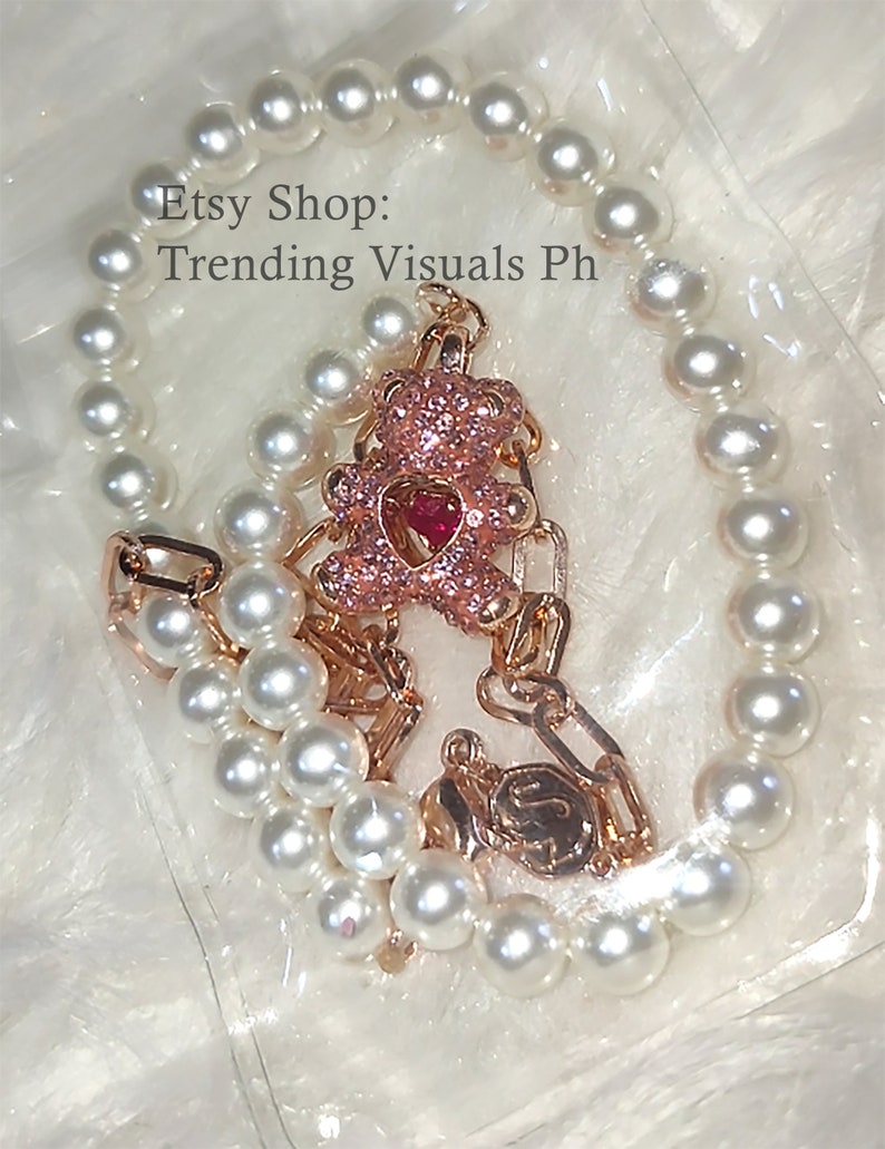 May include: A gold chain necklace with a pink sparkly teddy bear charm and a white pearl bracelet. The necklace and bracelet are on a white background. Etsy Shop: Trending Visuals Ph