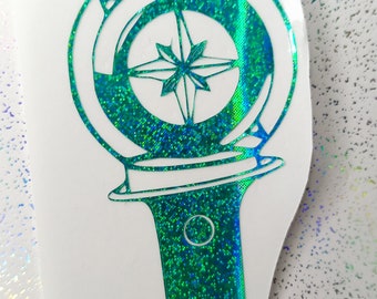 May include: A green and blue iridescent sticker of a light stick with a compass design. The compass has a star in the center and is surrounded by a circle.