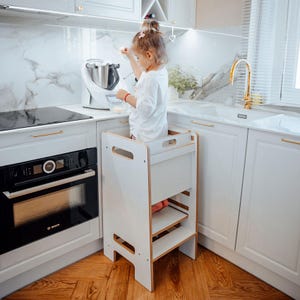 Può includere: Uno sgabello da cucina in legno bianco con due gradini e una piattaforma superiore. Lo sgabello è progettato per aiutare i bambini a raggiungere i piani di lavoro. Il bambino è in piedi sulla piattaforma, vicino a un elettrodomestico bianco.