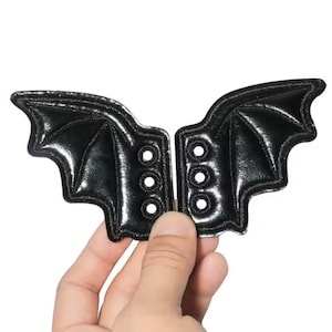 May include: Two black, bat-wing shaped patches with three sets of eyelets. The patches are held by a hand against a white background. The wings have a shiny, textured appearance.
