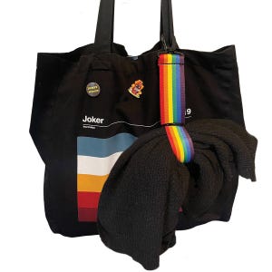 Può includere: Borsa tote nera con cinturino arcobaleno e maglione nero. La borsa presenta la parola "Joker" e una grafica colorata. Due spille sono attaccate alla borsa. Il cinturino è un vibrante arcobaleno.