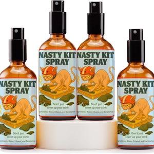 Op de afbeelding: Vier amberkleurige spuitflessen met het opschrift "NASTY KIT SPRAY". Elke fles heeft een cartoonkat met een helm en de slogan: "Don't just cover up your stink". De flessen staan op een wit platform.