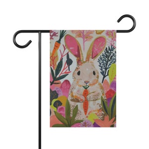 May include: A garden flag featuring a whimsical illustration of a bunny holding a carrot, surrounded by colorful foliage. The bunny has pink ears and is set against a backdrop of pink, orange, and green plants. The flag is mounted on a black metal stand.