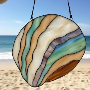 May include: A stained glass suncatcher in a rounded shape, with wavy bands of colour. The colours include shades of brown, beige, blue, and green, with black outlines. The suncatcher is suspended by a black cord, ideal for coastal or beach-themed decor.