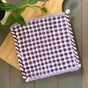 May include: A purple and white checkered potholder with white trim rests on a wooden cutting board. A small green plant in a white pot is in the upper left corner. The potholder is square and has a loop for hanging.
