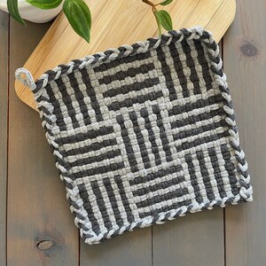 May include: A square, woven potholder in shades of gray. The design features a geometric pattern of alternating dark and light gray stripes. The edges are finished with a braided border and a small loop for hanging. This kitchen accessory is designed for heat resistance.