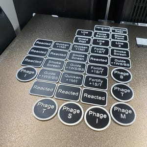 May include: A collection of black and silver game tokens and rectangular labels with white text. The labels include terms like "Reacted," "Quicken," and "Smoke Screen." The round tokens are labeled "Phage" with a letter.