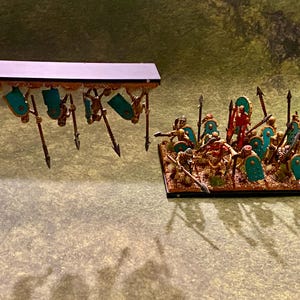 Puede incluir: Figuras en miniatura de una legi&oacute;n romana, con escudos turquesa, lanzas y detalles dorados. Las figuras est&aacute;n sobre una base rectangular, con una segunda unidad sostenida arriba. Ideal para juegos de mesa.
