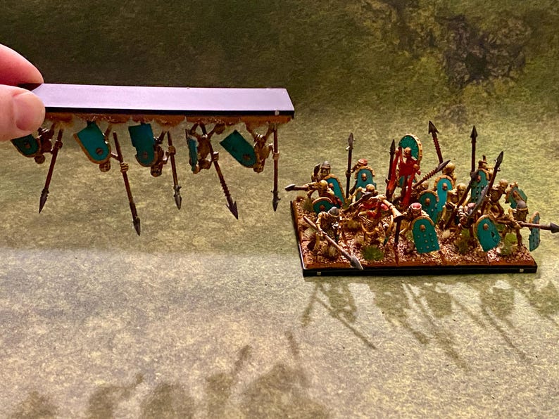 Puede incluir: Figuras en miniatura de una legi&oacute;n romana, con escudos turquesa, lanzas y detalles dorados. Las figuras est&aacute;n sobre una base rectangular, con una segunda unidad sostenida arriba. Ideal para juegos de mesa.