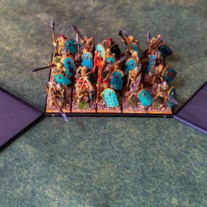 Puede incluir: Un ej&eacute;rcito en miniatura para un juego de mesa, compuesto por figuras con lanzas y escudos. Las figuras son doradas, rojas y turquesas. Dos bases rectangulares negras flanquean el ej&eacute;rcito sobre una superficie verde.