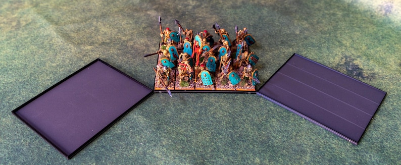 Puede incluir: Un ej&eacute;rcito en miniatura para un juego de mesa, compuesto por figuras con lanzas y escudos. Las figuras son doradas, rojas y turquesas. Dos bases rectangulares negras flanquean el ej&eacute;rcito sobre una superficie verde.