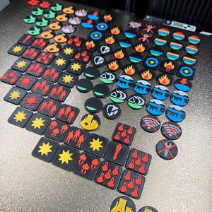 May include: A collection of 3D-printed game tokens and markers in various shapes and colors. The tokens include squares and circles with symbols like flames, people, and stars. Colors include red, yellow, green, blue, and black.