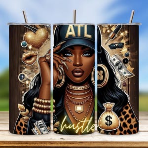 May include: A black and gold tumbler with a woman's face and the words "ATL Hustle" on it. The woman is wearing a gold chain, earrings, and a baseball cap that says "ATL".