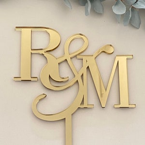 May include: Gold mirrored cake topper with the letters R & M, connected by a stylized ampersand. The topper is attached to a thin gold rod.