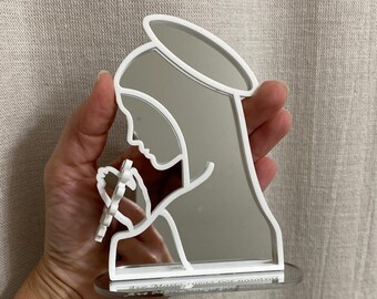 May include: A mirrored silhouette of a religious figure, featuring a halo and holding a pen. The figure is outlined in white, with a reflective surface. The base is clear, and the design is held in a hand.