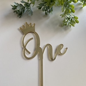 May include: A gold-colored cake topper with the word "One" in a script font, topped with a crown. The topper is designed to be inserted into a cake. The background is white with green foliage.