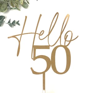 May include: Gold-colored cake topper with the words "Hello 50" in a cursive font and the number 50. The topper is held by a hand against a white background, with greenery in the upper left corner.