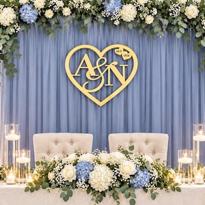 May include: A wedding sweetheart table setup with a white sequin tablecloth, two tufted chairs, and a floral centerpiece of white roses, blue hydrangeas, and eucalyptus. A gold heart-shaped sign with the letters A&N and the date 04.16.2026 hangs above.