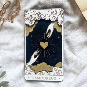 Puede incluir: Una carta de tarot de madera con fondo negro, un corazón dorado y líneas radiantes. Dos manos blancas se extienden hacia el corazón. La carta está enmarcada con nubes doradas y el texto "L'AMOUREUX" en la parte inferior.