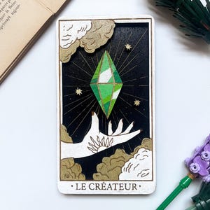 以下が含まれることがあります： 緑色の宝石、白い手、金色のアクセントが施された木製のタロットカード。カードの下部には「LE CRÉATEUR」の文字。デザインには星と雲のような形が含まれています。