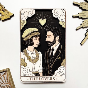 May include: Wooden tarot card featuring a couple, with a heart and sunburst design. The card has a black background with gold accents and the words "THE LOVERS" at the bottom. The card is surrounded by white clouds.