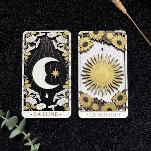 Peut inclure: Deux cartes de tarot en bois. L'une représente un croissant de lune avec un visage, une étoile et le texte "LA LUNE". L'autre carte montre un soleil doré entouré de tournesols et le texte "LE SOLEIL".