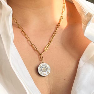 Könnte beinhalten: Goldfarbene Kette mit rundem Anhänger. Der Anhänger hat ein weißes und schwarzes Design mit kleinen, runden Verzierungen und einem zentralen, strukturierten Element. Die Kette wird über einem weißen Hemd getragen.