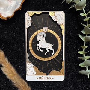 Peut inclure: Une carte rectangulaire avec une illustration de bélier blanc dans un cercle zodiacal doré sur fond noir. La carte a des bordures décoratives dorées et blanches, avec le mot "BÉLIER" en bas.