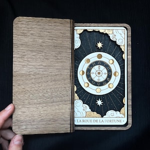 May include: Wooden tarot card box with a card featuring a Wheel of Fortune design. The card has a white and gold circular design with astrological symbols, set against a dark blue background with stars and clouds. The text "LA ROUE DE LA FORTUNE" is at the bottom.
