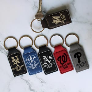 May include: A collection of personalized keychains in various colors, including black, blue, red, and gray. Each keychain features a team logo and the text "YOUR TEXT HERE". A key is attached to a brown keychain with the letter "M".