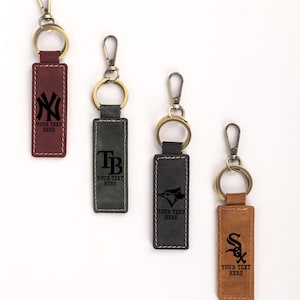 May include: Four leather keychains with metal hardware. Each keychain has a rectangular leather tag with a team logo and the text "YOUR TEXT HERE". The keychains are in burgundy, green, black, and tan.