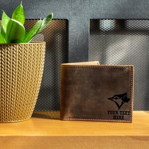 May include: A brown leather wallet with a stitched border. The wallet features a black graphic of a bird and the text "YOUR TEXT HERE". The wallet is displayed on a wooden surface.