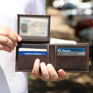May include: Open brown leather wallet with multiple card slots and a clear ID window. The wallet contains credit cards and cash. The wallet is held open, displaying its contents.
