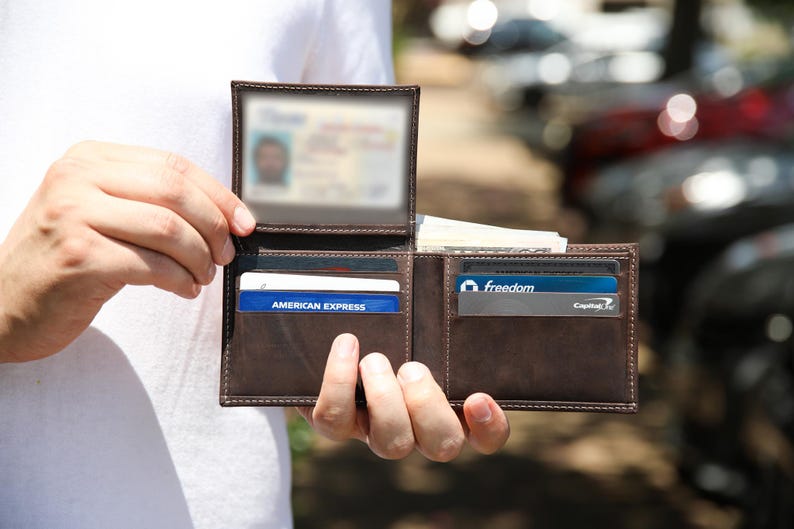 May include: Open brown leather wallet with multiple card slots and a clear ID window. The wallet contains credit cards and cash. The wallet is held open, displaying its contents.