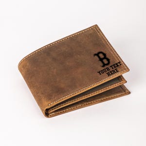 May include: Brown leather wallet with a stitched border. The wallet features a black letter "B" logo and the text "YOUR TEXT HERE". The wallet is folded and appears to be new.