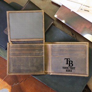 May include: Open brown leather wallet with multiple card slots and a bill compartment. The wallet has the initials "TB" and the text "YOUR TEXT HERE" engraved on the inside. Several other leather wallets are visible in the background.