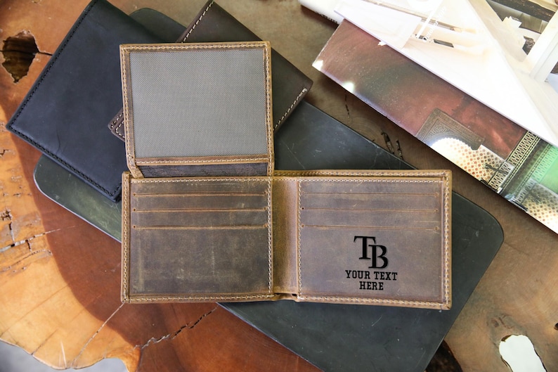 May include: Open brown leather wallet with multiple card slots and a bill compartment. The wallet has the initials "TB" and the text "YOUR TEXT HERE" engraved on the inside. Several other leather wallets are visible in the background.