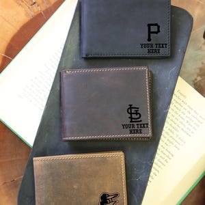 May include: Three leather wallets in black, brown, and tan, each with a different team logo and the text "YOUR TEXT HERE". The wallets are displayed on a dark surface with an open book.