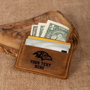 May include: A brown leather wallet with a black Baltimore Ravens logo and the text "YOUR TEXT HERE". The wallet is open and has a few dollar bills and credit cards inside.