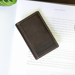 Pu&ograve; includere: Un portafoglio in pelle marrone con un bordo cucito e pi&ugrave; slot per carte. Il portafoglio &egrave; su una superficie bianca con un libro e una pianta sullo sfondo.
