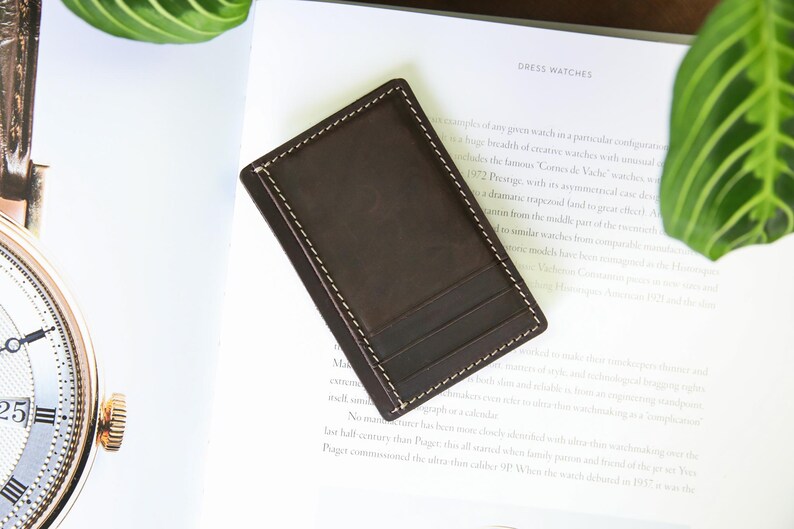 Pu&ograve; includere: Un portafoglio in pelle marrone con un bordo cucito e pi&ugrave; slot per carte. Il portafoglio &egrave; su una superficie bianca con un libro e una pianta sullo sfondo.