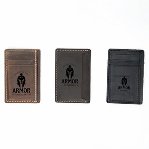 Pu&ograve; includere: Tre portafogli in pelle con un design minimalista. Ogni portafoglio presenta un logo nero con un elmo spartano e il testo "Armor Experiences". I portafogli sono marroni, marrone scuro e neri.