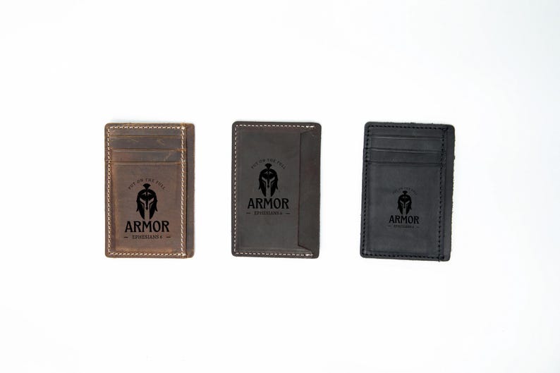 Pu&ograve; includere: Tre portafogli in pelle con un design minimalista. Ogni portafoglio presenta un logo nero con un elmo spartano e il testo "Armor Experiences". I portafogli sono marroni, marrone scuro e neri.