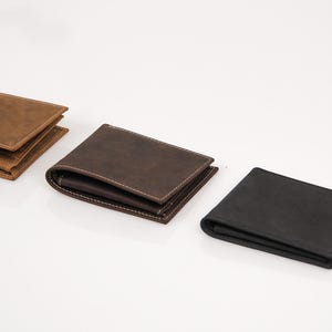 May include: Three leather wallets in different colors are displayed on a white surface. The wallets are brown, dark brown, and black. The brown wallet is on the left, the dark brown wallet is in the middle, and the black wallet is on the right.