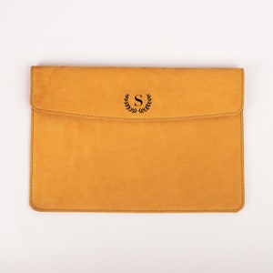 May include: Mustard-colored leather laptop sleeve with a flap closure. The sleeve features a black stitched border and a black monogram with the letter "S" inside a laurel wreath. The background is white.