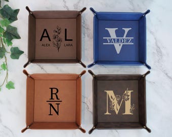 May include: Four personalized leather trays with different designs. The top left tray is brown with the initials "A & L" and the names "Alex & Lara" underneath. The top right tray is blue with the initial "V" and the name "Valdez" underneath. The bottom left tray is brown with the initials "R & N" stacked on top of each other. The bottom right tray is brown with the initial "M" and a floral design around it.