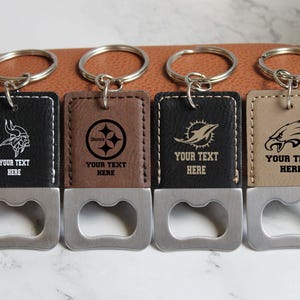 May include: Four different NFL team logo bottle openers with keychains. The bottle openers are made of metal and have a brown leatherette finish. Each bottle opener has a different team logo: Minnesota Vikings, Pittsburgh Steelers, Miami Dolphins, and Philadelphia Eagles. The bottle openers have the text "Your Text Here" engraved on them.
