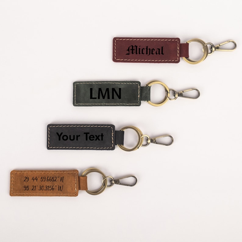 K&ouml;nnte beinhalten: Vier Leder-Schl&uuml;sselanh&auml;nger mit verschiedenen Gravuren. Die Schl&uuml;sselanh&auml;nger sind braun, gr&uuml;n, schwarz und beige. Die Gravuren beinhalten "Micheal", "LMN", "Your Text" und Koordinaten.